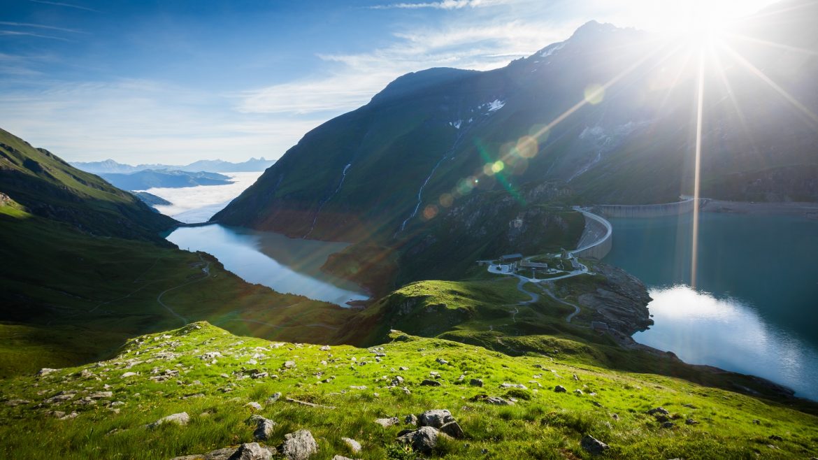Sonnenaufgang über einem Bergsee, mit grünen Hügeln und einer kurvenreichen Straße am Wasser.