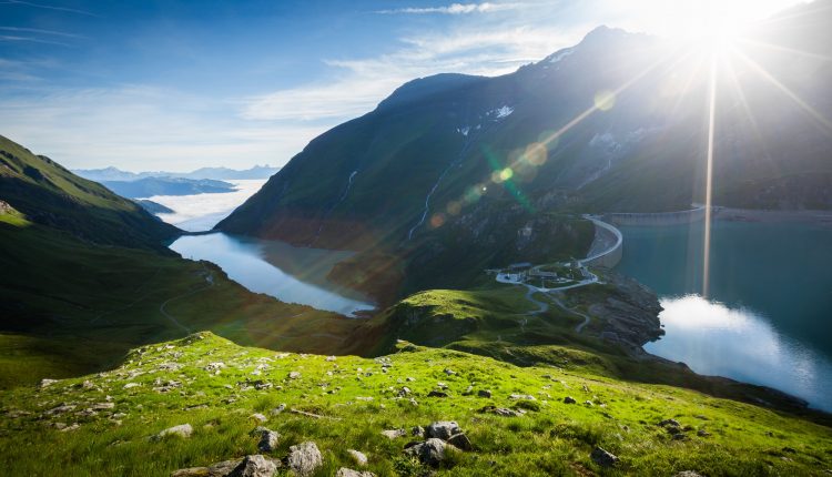 Sonnenaufgang über einem Bergsee, mit grünen Hügeln und einer kurvenreichen Straße am Wasser.