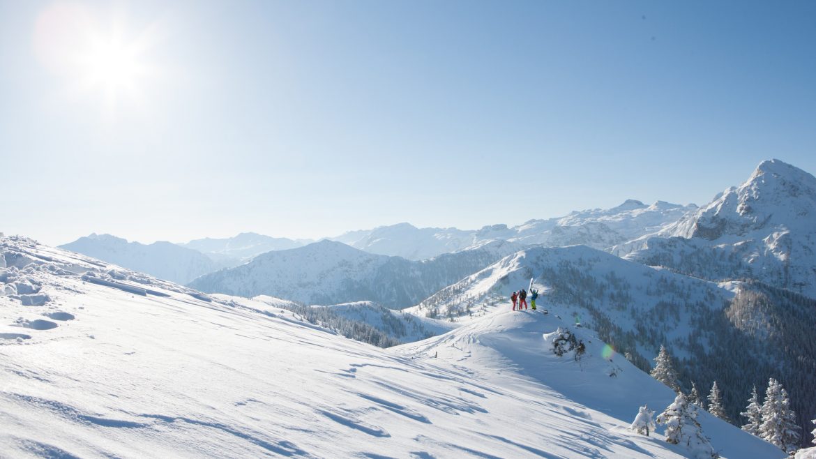 Menschen stehen auf einem verschneiten Bergrücken in der strahlenden Sonne mit Bergketten im Hintergrund.