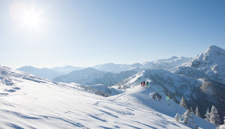 Menschen stehen auf einem verschneiten Bergrücken in der strahlenden Sonne mit Bergketten im Hintergrund.