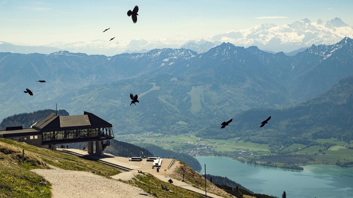 Berglandschaft mit einem Dorf am Seeufer, fliegenden Vögeln und einem Holzgebäude auf einem Hügel im Vordergrund.