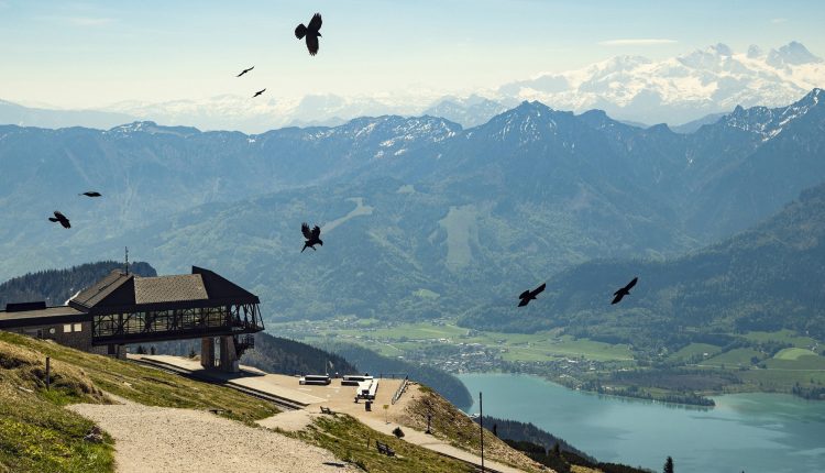 Berglandschaft mit einem Dorf am Seeufer, fliegenden Vögeln und einem Holzgebäude auf einem Hügel im Vordergrund.