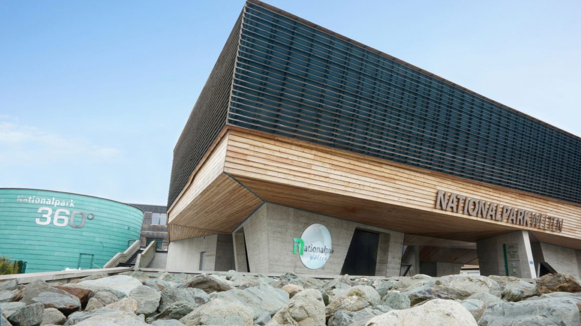 Modernes Gebäude mit Holz- und Glasfassade mit der Aufschrift "Nationalpark" neben einer runden Grünanlage und Felsen.