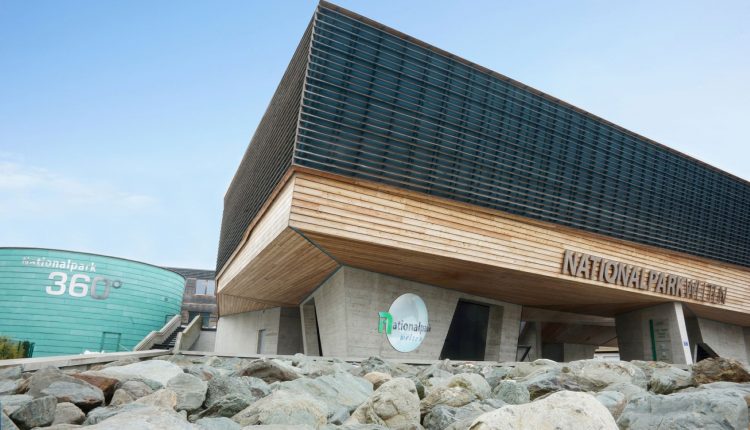 Modernes Gebäude mit Holz- und Glasfassade mit der Aufschrift "Nationalpark" neben einer runden Grünanlage und Felsen.