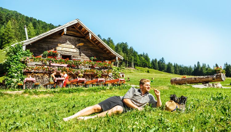 Ein Mann entspannt sich im Gras neben einer rustikalen, blumengeschmückten Hütte, in der Menschen an Tischen im Freien sitzen.